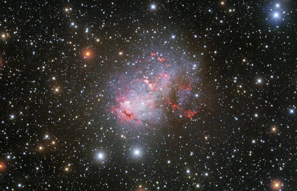 Dense star field with a bright, irregular starburst galaxy glowing pink and blue at center, surrounded by scattered stars.