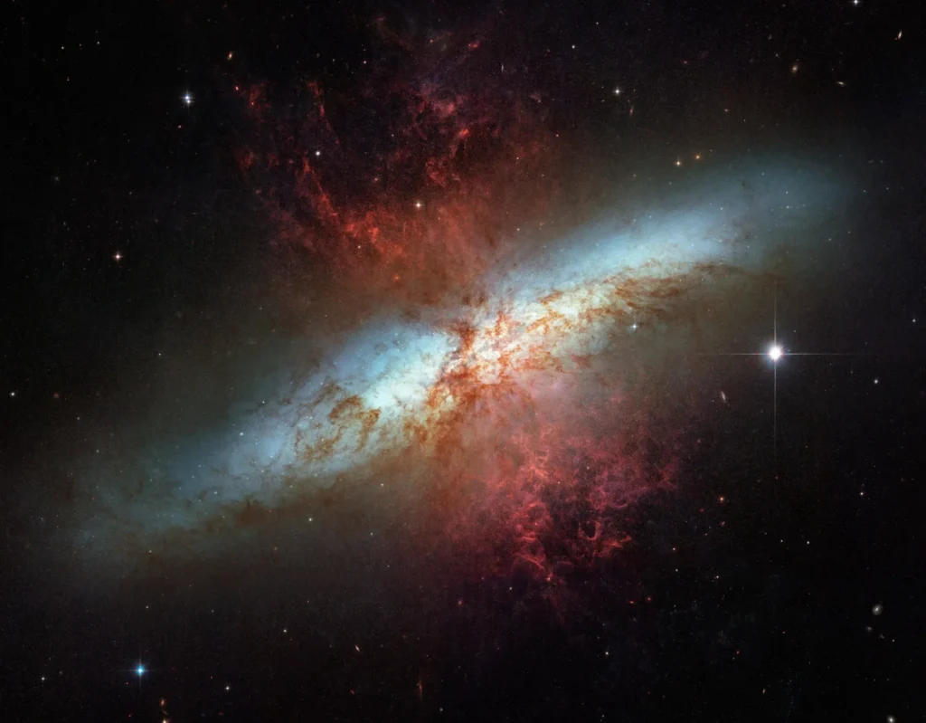 Starburst galaxy M82 with bright core, blue disk, and red gas plumes, surrounded by stars in deep space.