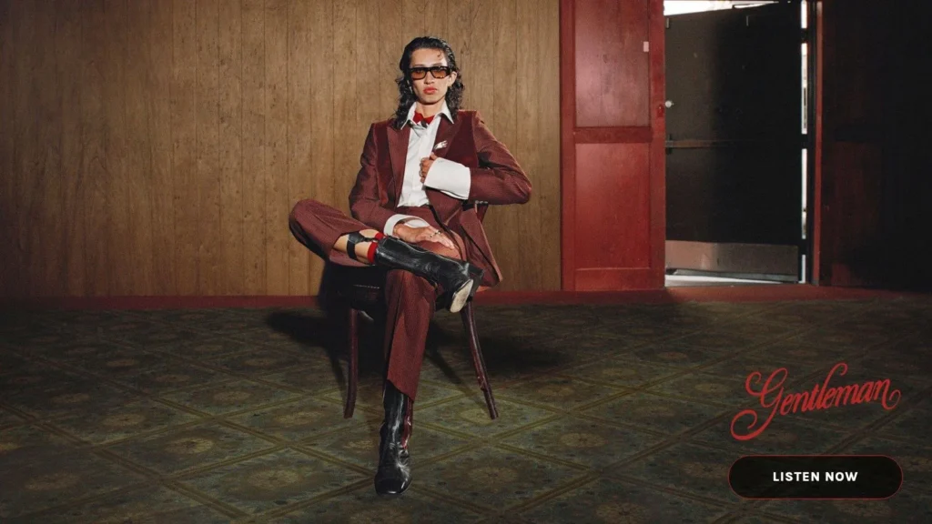Person in burgundy suit and sunglasses seated on chair in wood-paneled room, “Gentleman” text and “Listen Now” button.