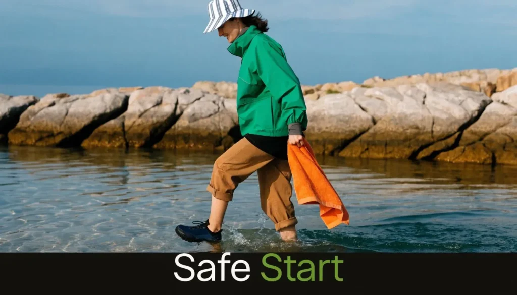 Person wading into cold seawater with towel and “Safe Start” text about beginner cold water swimming safety.