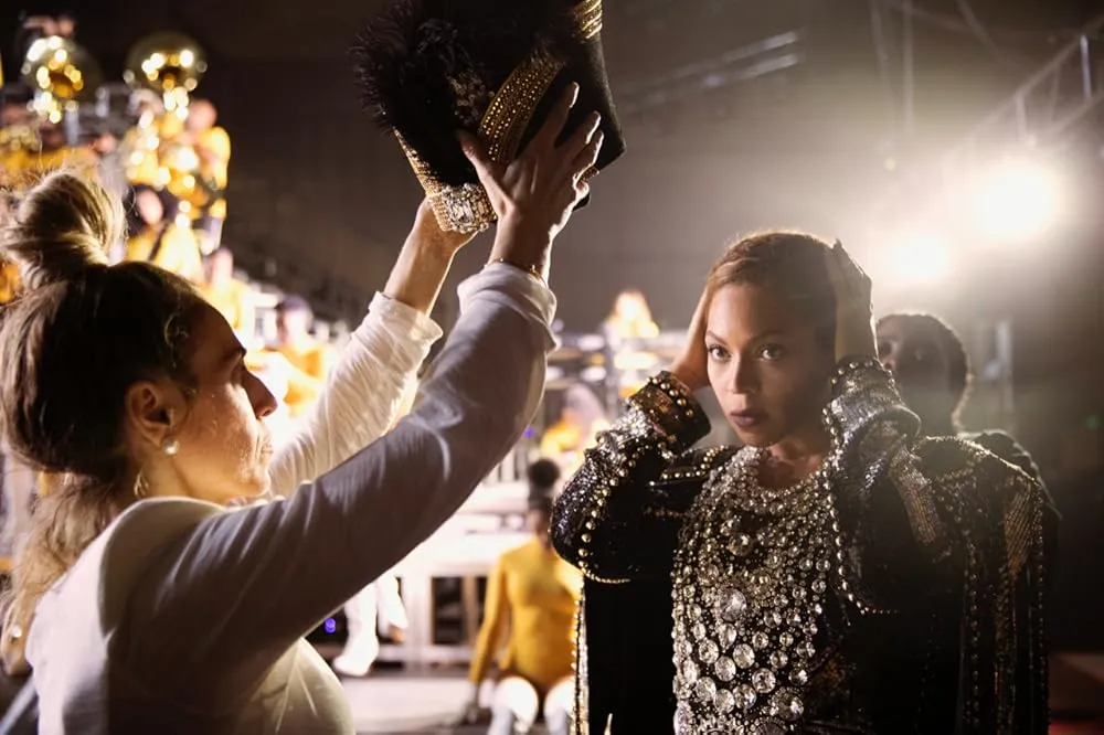 Performer in sparkling costume backstage while an assistant adjusts a decorative headpiece under bright stage lights.