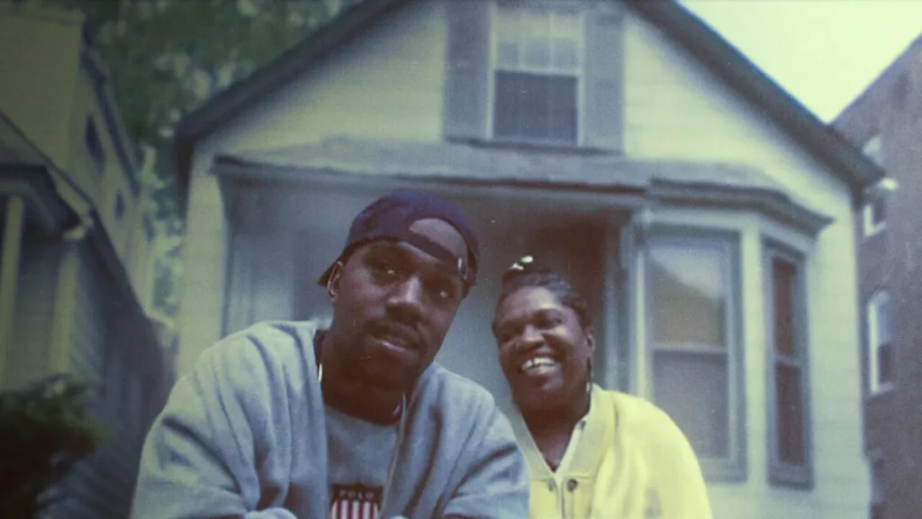 Two people smiling outside a house in a grainy vintage-style photo with a nostalgic documentary look.